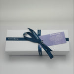 White gift box with a blue ribbon and label on a light gray background. Inside products include bath bomb, candle and soap. 