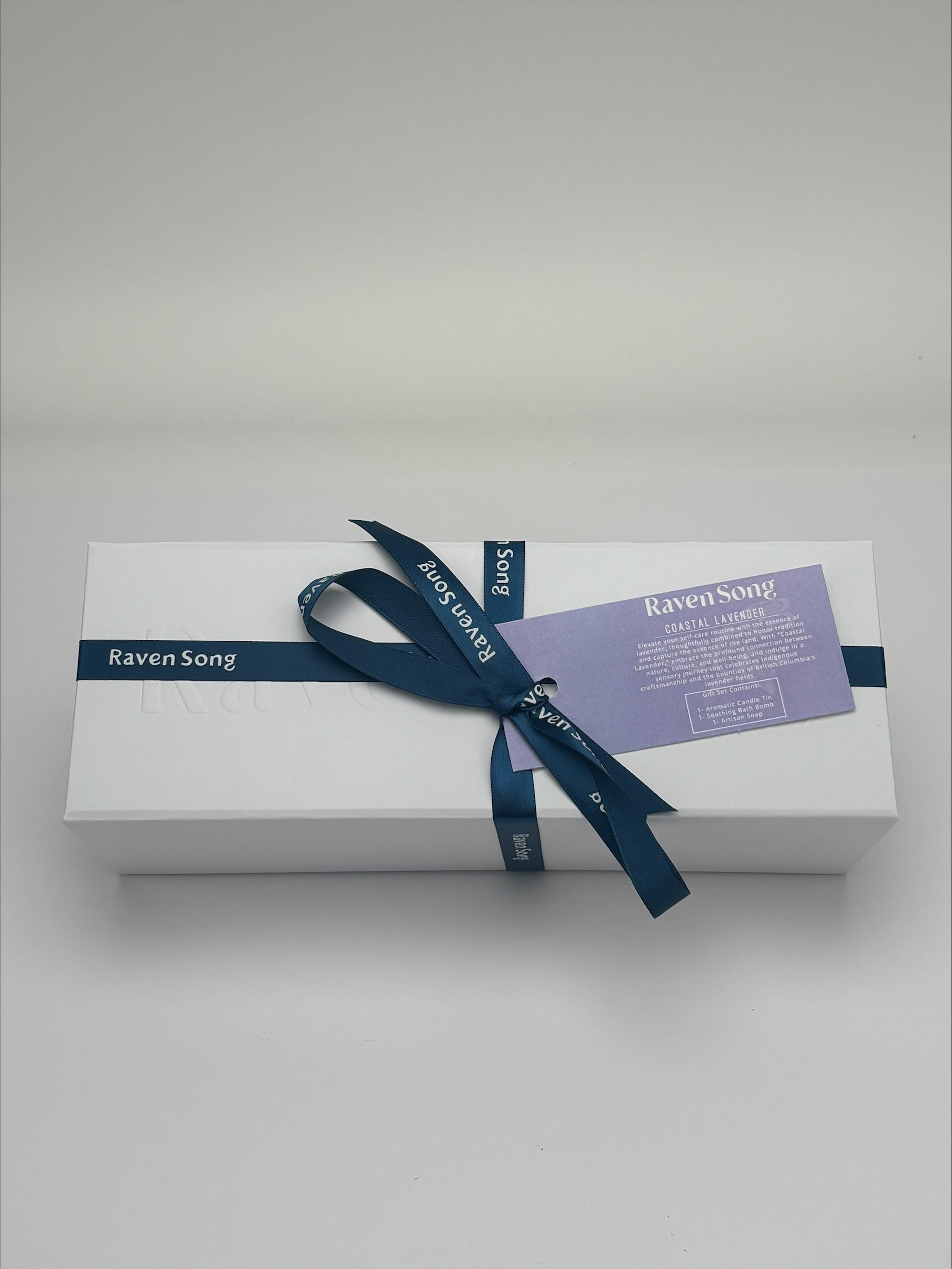White gift box with a blue ribbon and label on a light gray background. Inside products include bath bomb, candle and soap. 