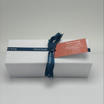 White gift box with a blue ribbon and label on a light gray background. Inside products include bath bomb, candle and soap. 