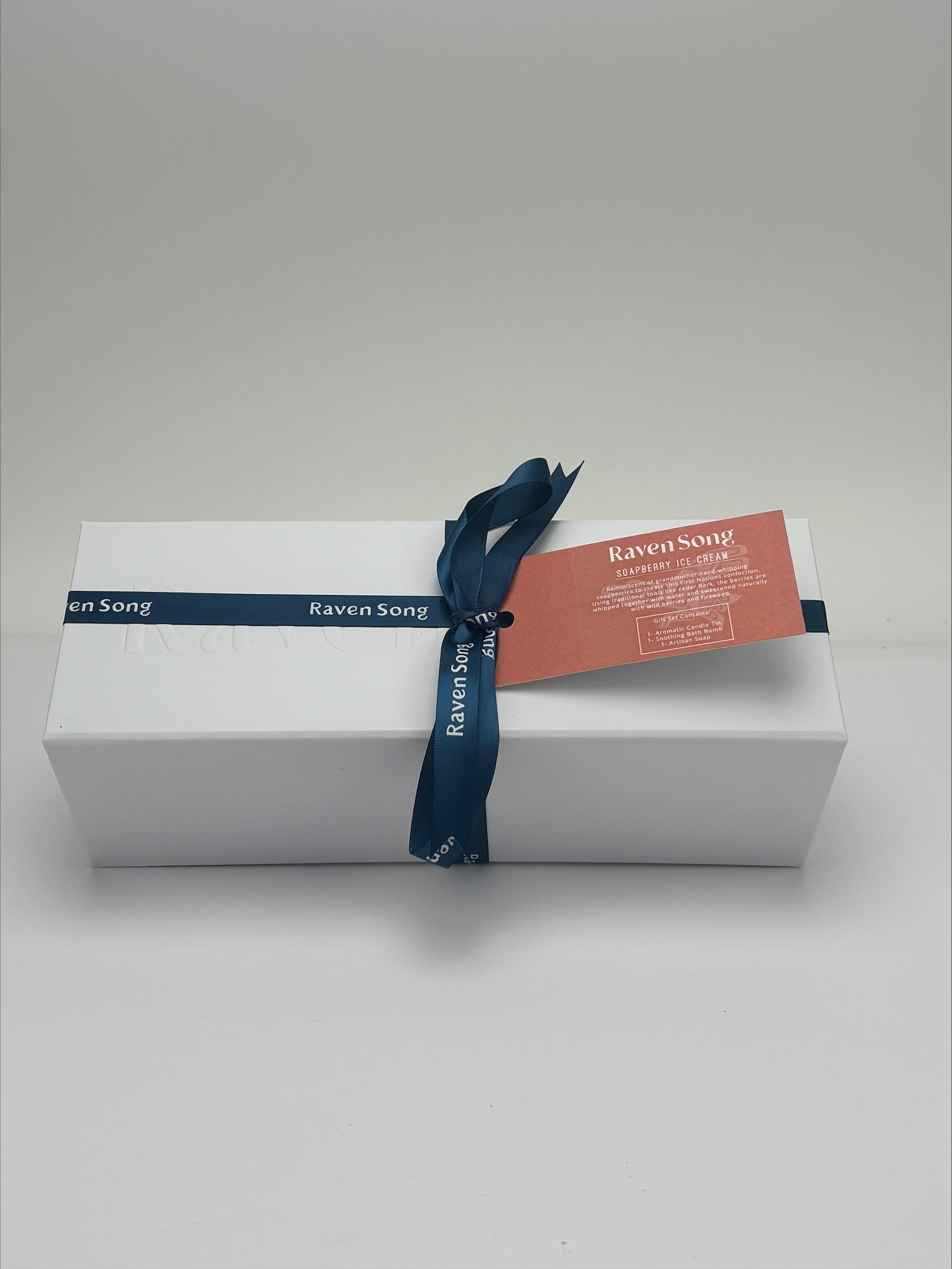 White gift box with a blue ribbon and label on a light gray background. Inside products include bath bomb, candle and soap. 